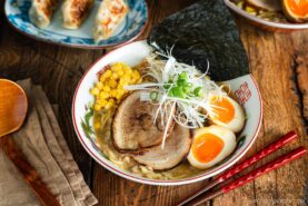 A bowl of ramen with sliced pork, soft-boiled eggs, corn, green onions, seaweed, and noodles in broth. Red chopsticks and a wooden spoon are nearby. Gyoza dumplings are in the background on a wooden table.