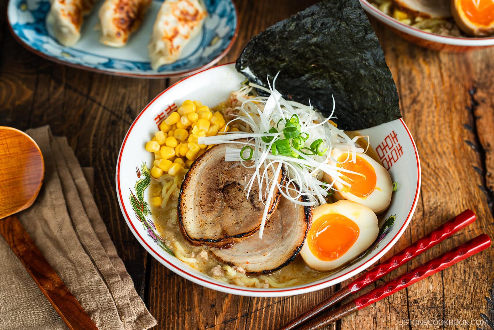 A bowl of ramen with sliced pork, soft-boiled eggs, corn, green onions, seaweed, and noodles in broth. Red chopsticks and a wooden spoon are nearby. Gyoza dumplings are in the background on a wooden table.