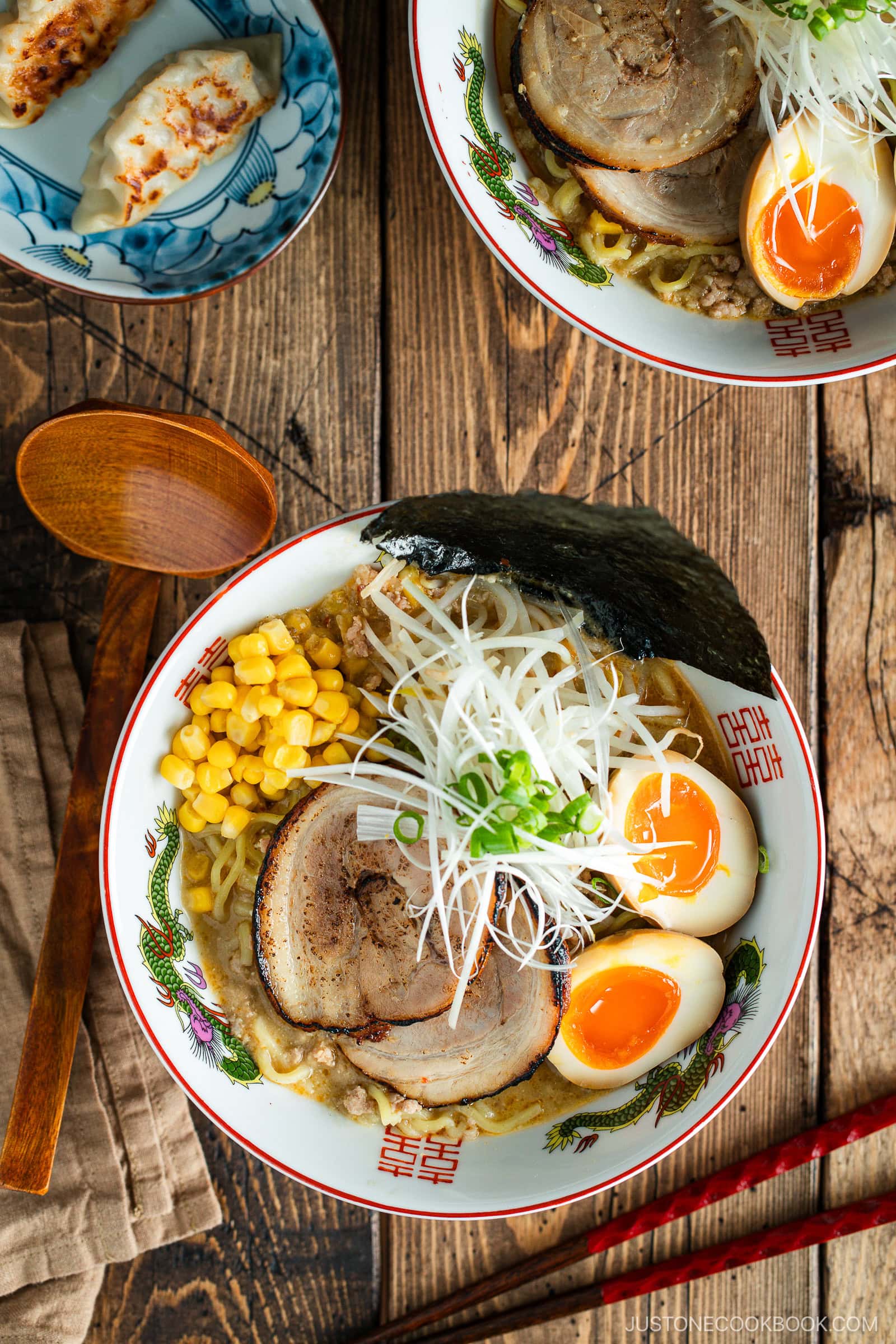 A bowl of ramen topped with sliced pork, corn, soft-boiled eggs, nori, and shredded green onions, next to a plate of gyoza dumplings on a rustic wooden table with chopsticks and a spoon.