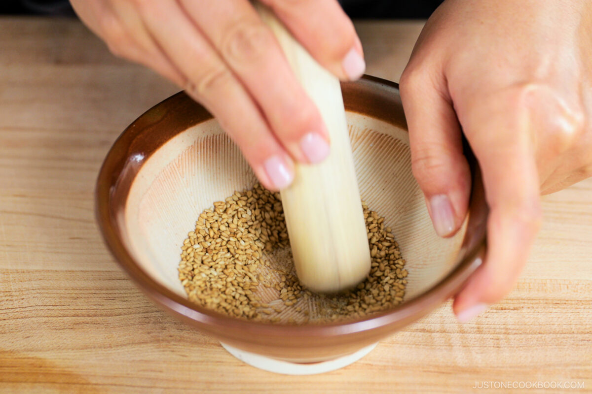 Close-up of hands using a wooden pestle to grind sesame seeds in a small brown and white bowl on a wooden surface.
