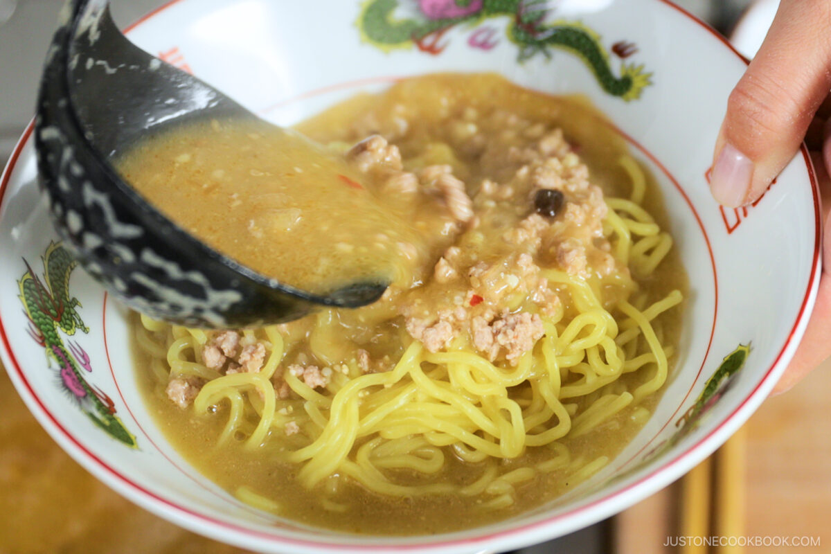 A close-up of a bowl with yellow noodles topped with minced meat, as a ladle pours hot, creamy broth over them. The bowl features colorful dragon designs on the rim.