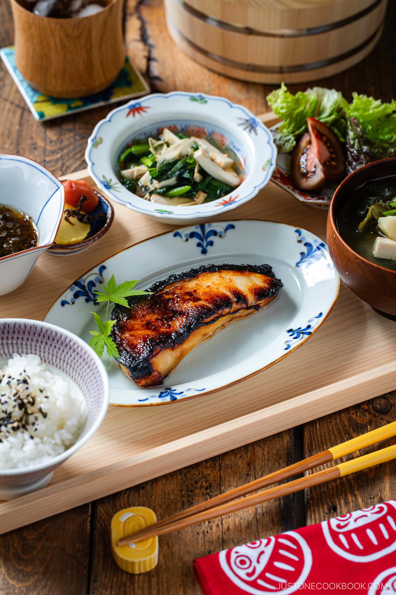 A Japanese meal set on a wooden tray features grilled fish, rice, miso soup, a vegetable side dish, and leafy greens, with chopsticks and a red napkin nearby. The dishes are arranged neatly in small bowls and plates.