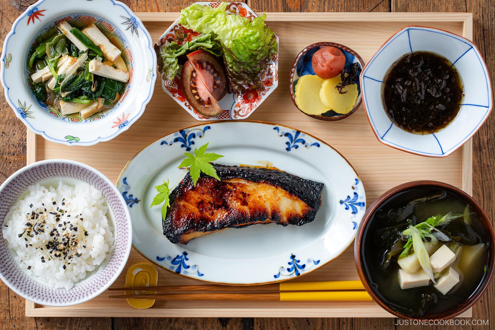 A Japanese breakfast set with grilled fish, rice, miso soup with tofu, pickles, greens, seaweed salad, and a side salad with tomato and lettuce, all neatly arranged on a wooden tray.