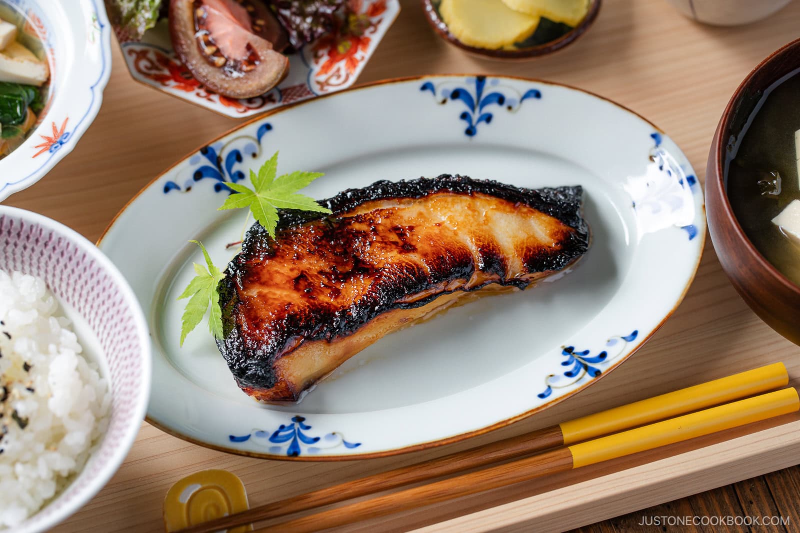 A plate with a glazed, grilled fish fillet garnished with a green leaf, surrounded by bowls of rice, pickles, soup, and salad, with chopsticks resting nearby on a wooden table.