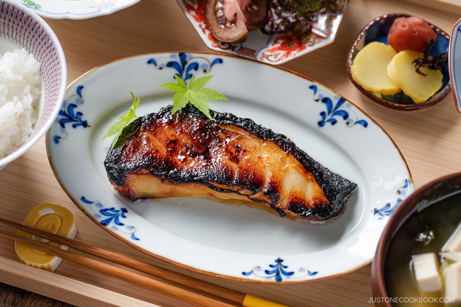 A grilled, caramelized fish fillet garnished with green leaves is served on a decorative plate, surrounded by rice, pickles, soup, and side dishes on a wooden tray.
