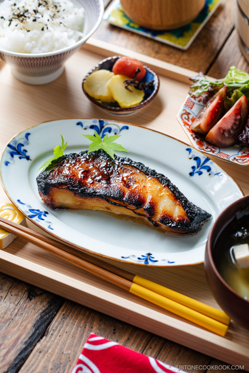 A Japanese meal with glazed grilled fish on a decorative plate, steamed rice, pickled vegetables, fresh salad, and miso soup, served on a wooden tray with chopsticks.