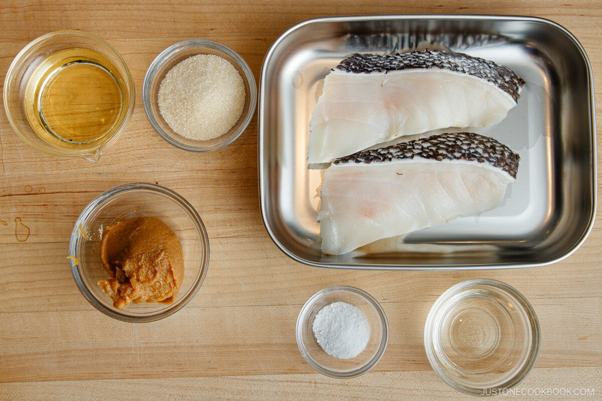 Raw fish fillets on a metal tray, surrounded by small bowls containing miso paste, sugar, salt, sake, mirin, and a clear liquid, all on a wooden surface.