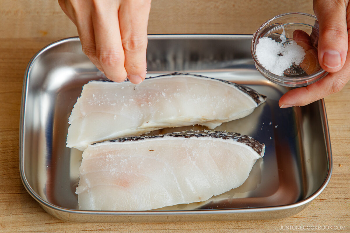 A hand sprinkles salt from a small glass bowl onto two raw fish fillets placed in a metal tray on a wooden surface.