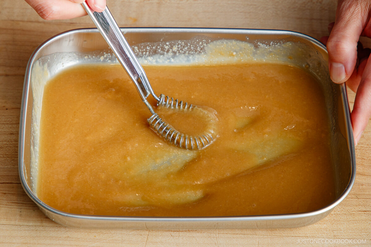 A person uses a whisk to mix a thick, light brown sauce or batter in a rectangular metal tray on a wooden surface.
