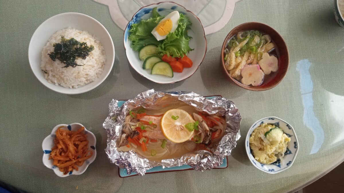 A Japanese meal set on a table, featuring white rice, salad with boiled egg, miso soup, grilled fish with lemon in foil, seasoned vegetables, and a small dish of potato salad.