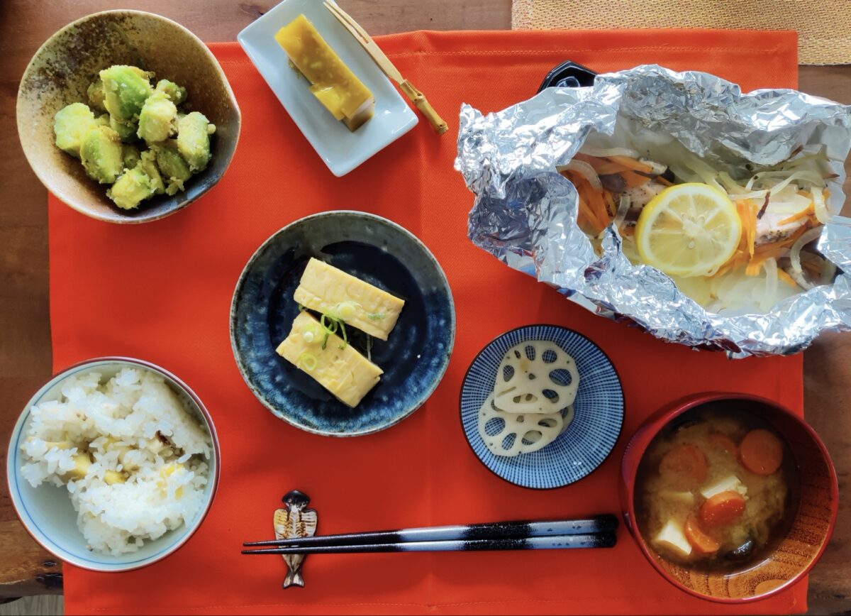 A Japanese meal on a red placemat with rice, miso soup, rolled omelet, avocado salad, lotus root slices, and fish with lemon and vegetables in foil. Chopsticks and a chopstick rest are placed in the center.
