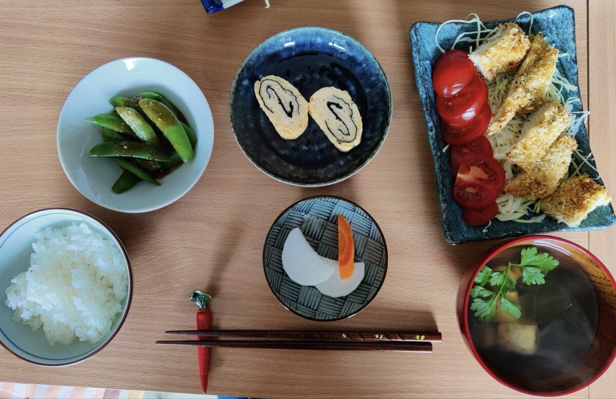 A Japanese meal set on a wooden table includes rice, miso soup, sliced cucumber, tamagoyaki, pickled vegetables, sliced tomatoes, and breaded fried fish. Chopsticks rest in the center.