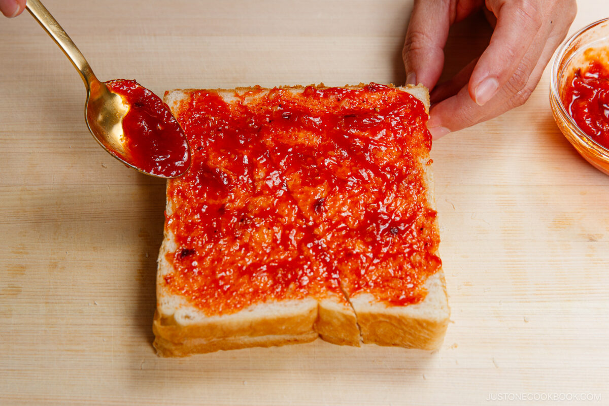 A hand spreads bright red jam onto thick slices of white bread with a spoon on a wooden surface. A small bowl of jam is visible to the side.