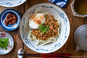 A bowl of udon noodles topped with minced meat, a poached egg, sliced green onions, and seasoning, surrounded by small dishes of chili oil, more green onions, and tea on a wooden table.