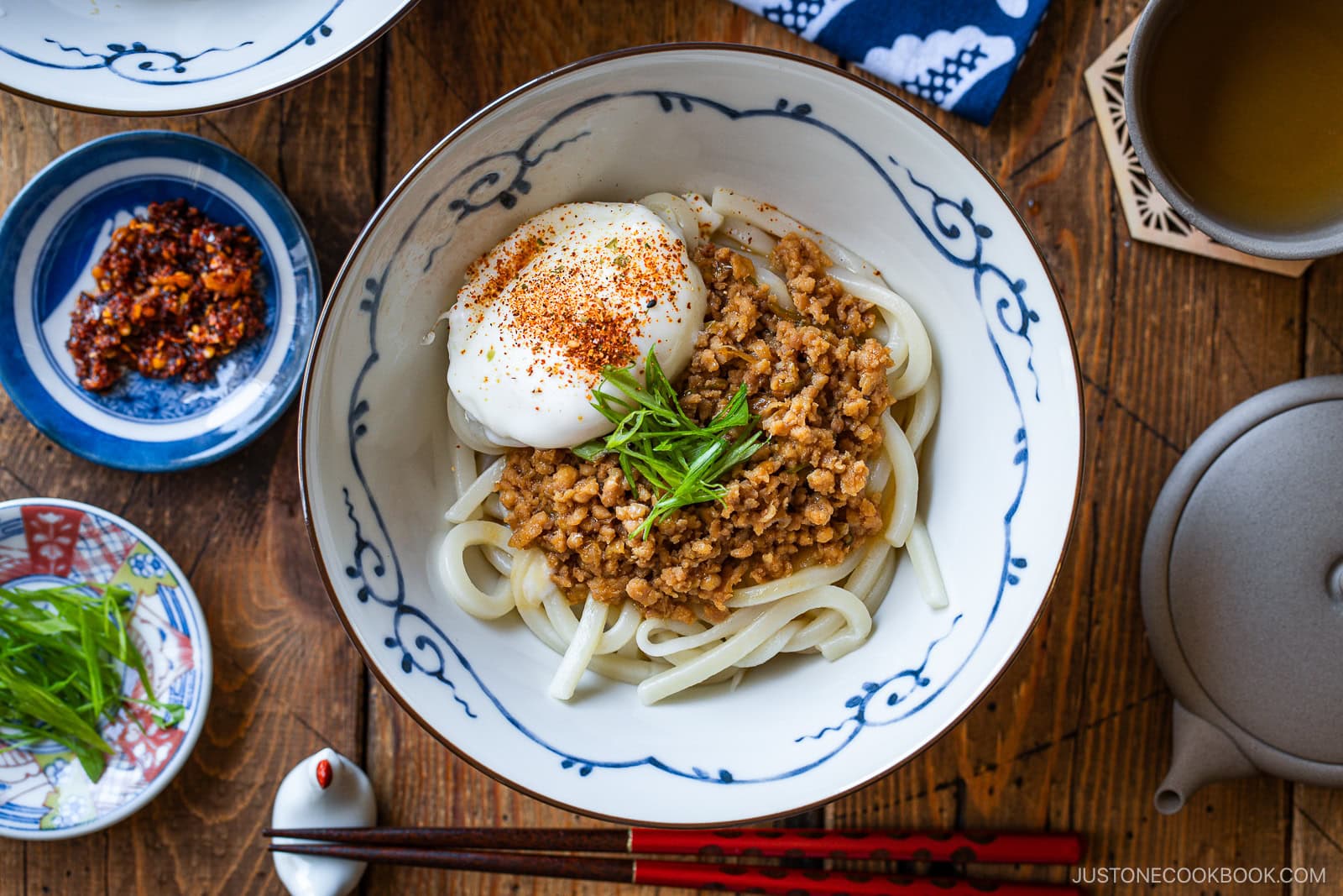 A bowl of udon noodles topped with minced meat, a poached egg, sliced green onions, and seasoning, surrounded by small dishes of chili oil, more green onions, and tea on a wooden table.