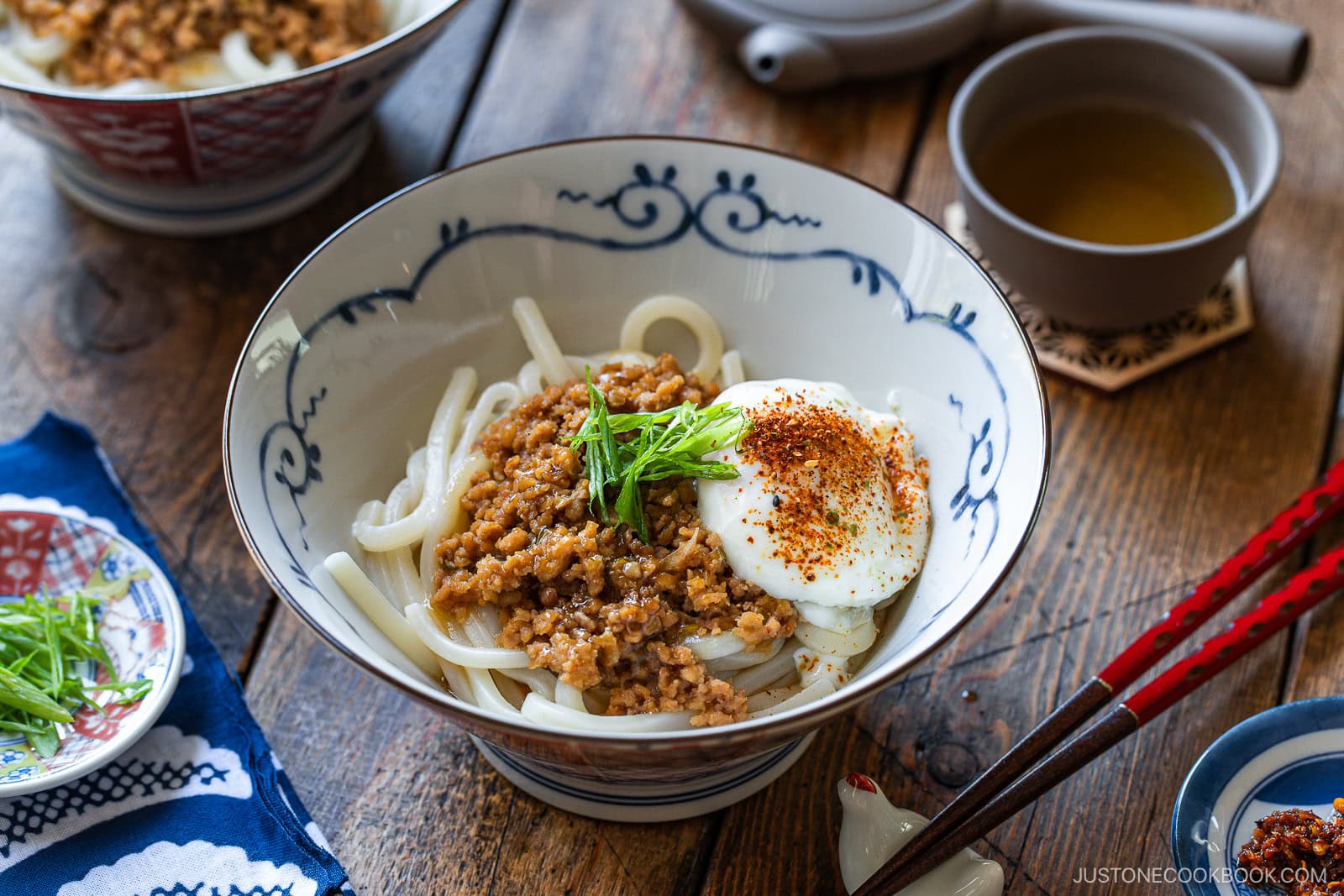 A bowl of udon noodles topped with minced meat, a soft-boiled egg, sliced green onions, and red seasoning, accompanied by chopsticks and a cup of tea on a wooden table.