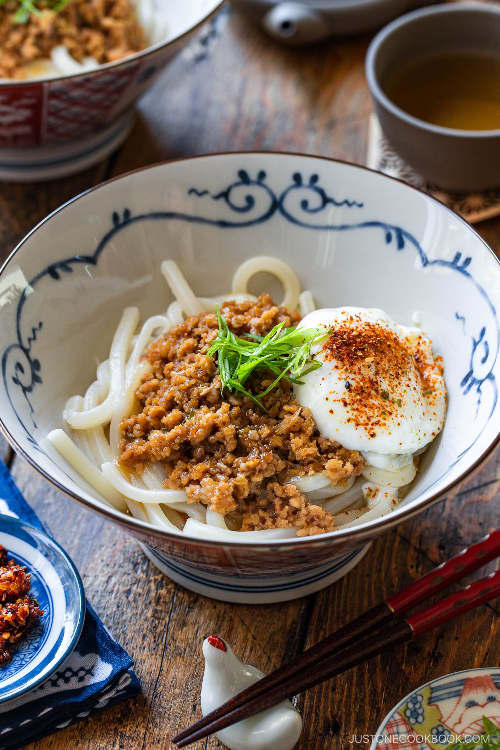 A bowl of udon noodles topped with seasoned ground meat, a soft-boiled egg, sliced green onions, and red pepper flakes, served on a wooden table with tea and condiments nearby.