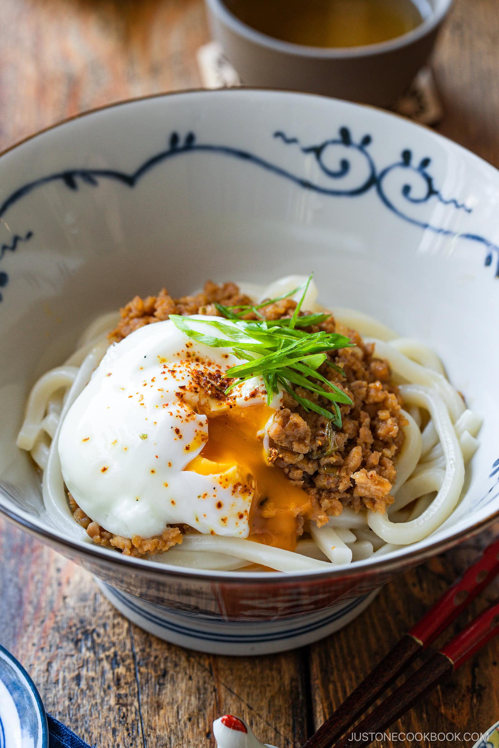 A bowl of udon noodles topped with seasoned ground meat, a poached egg with a runny yolk, sliced green onions, and a sprinkle of spices, served in a decorative bowl on a wooden table.