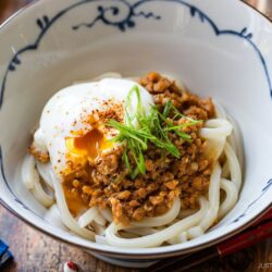 A bowl of udon noodles topped with minced meat, a soft-boiled egg with a runny yolk, sliced green onions, and a sprinkle of spices, served in a decorative white and blue bowl.