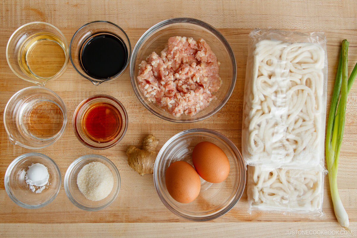 Top-down view of various ingredients on a wooden surface, including ground meat, frozen udon noodles, eggs, green onion, ginger, and small bowls of sauces, oil, seasonings, and cornstarch.