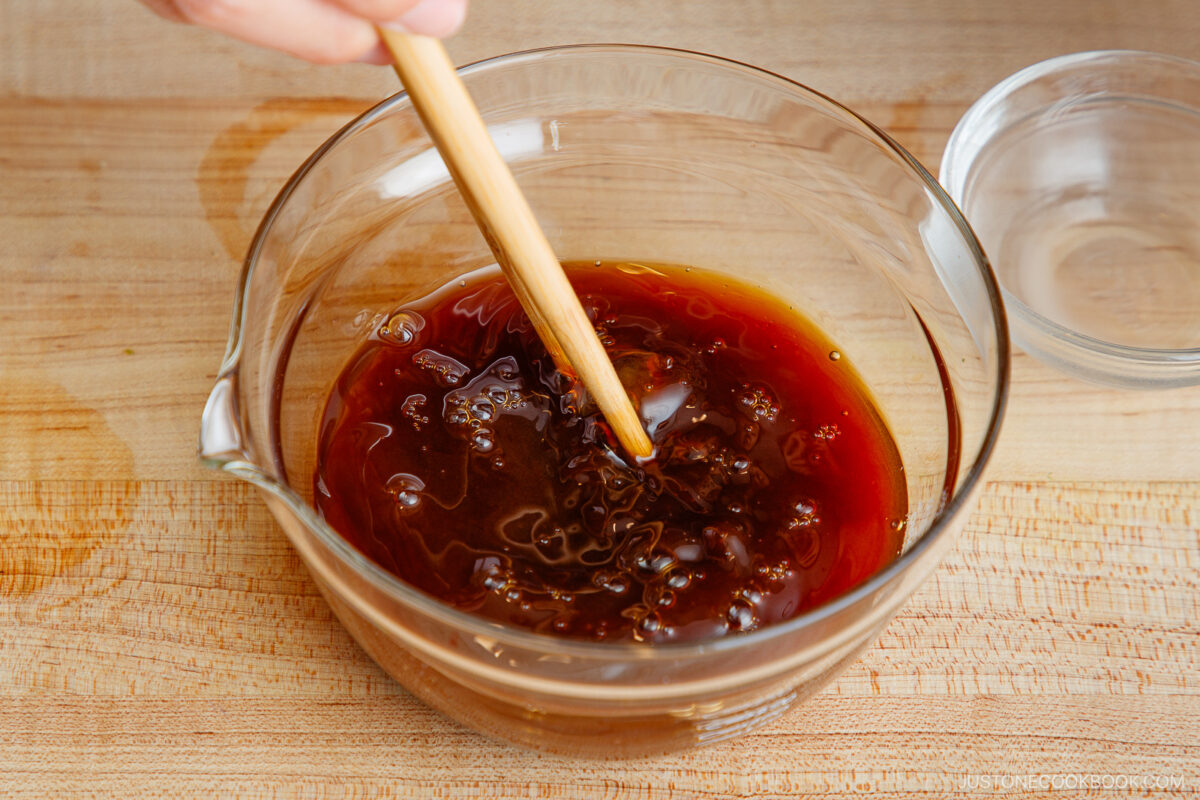 A person stirs a dark brown liquid mixture in a clear glass bowl with a wooden chopstick on a wooden countertop.