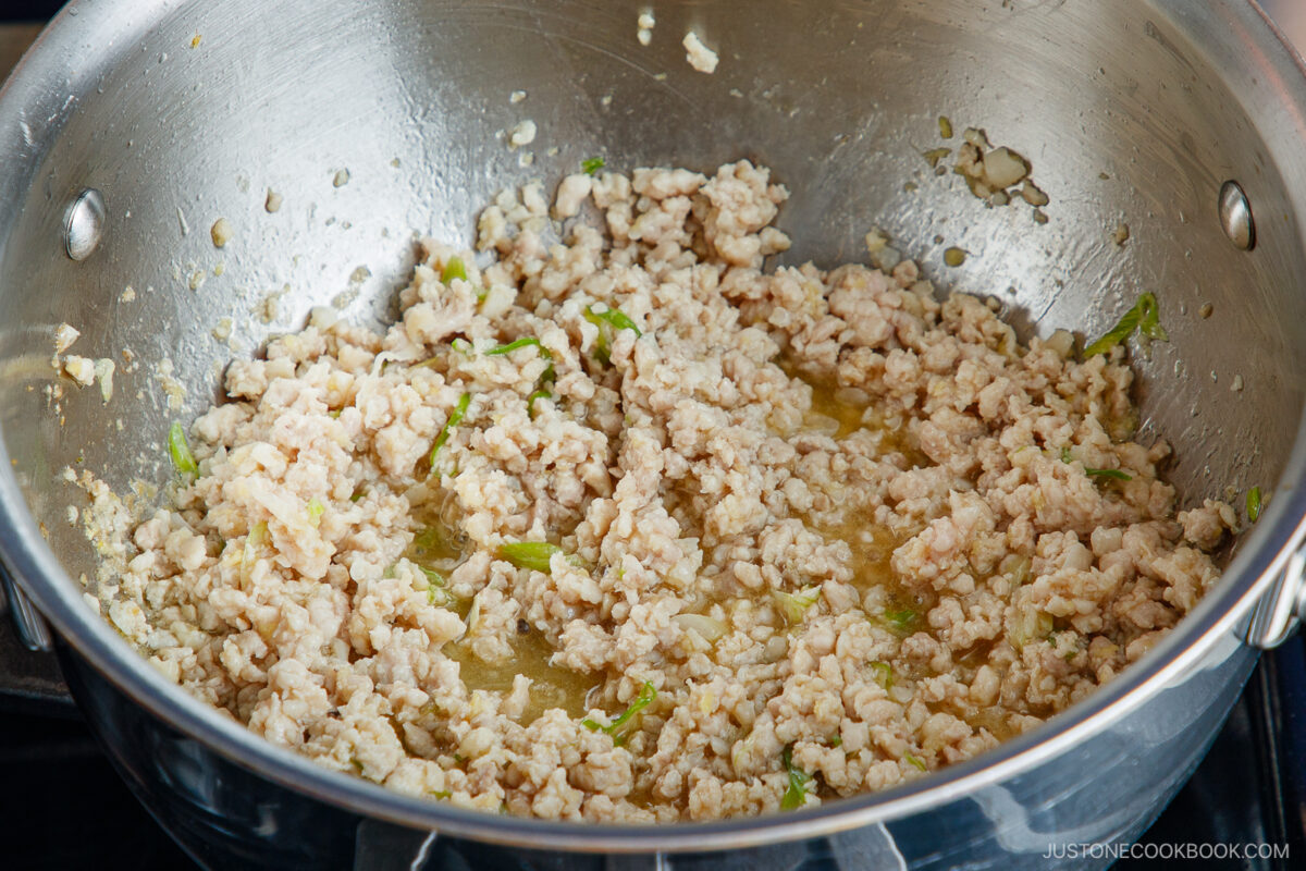 Ground chicken being cooked with chopped green onions in a stainless steel pan, with some liquid visible at the bottom. The mixture appears lightly browned and is being prepared on a stovetop.