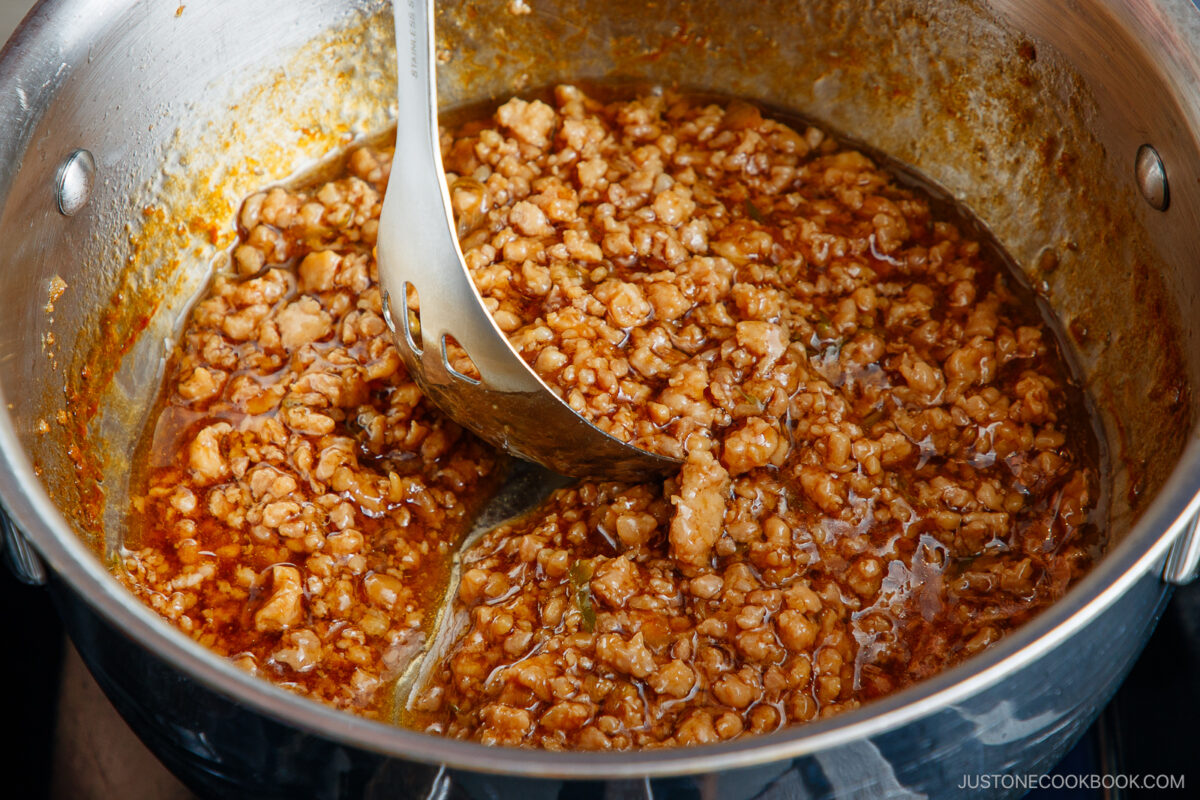 Ground meat cooked in a rich, reddish-brown sauce, being stirred with a slotted spoon inside a stainless steel pot. The sauce looks thick and savory with visible oil.