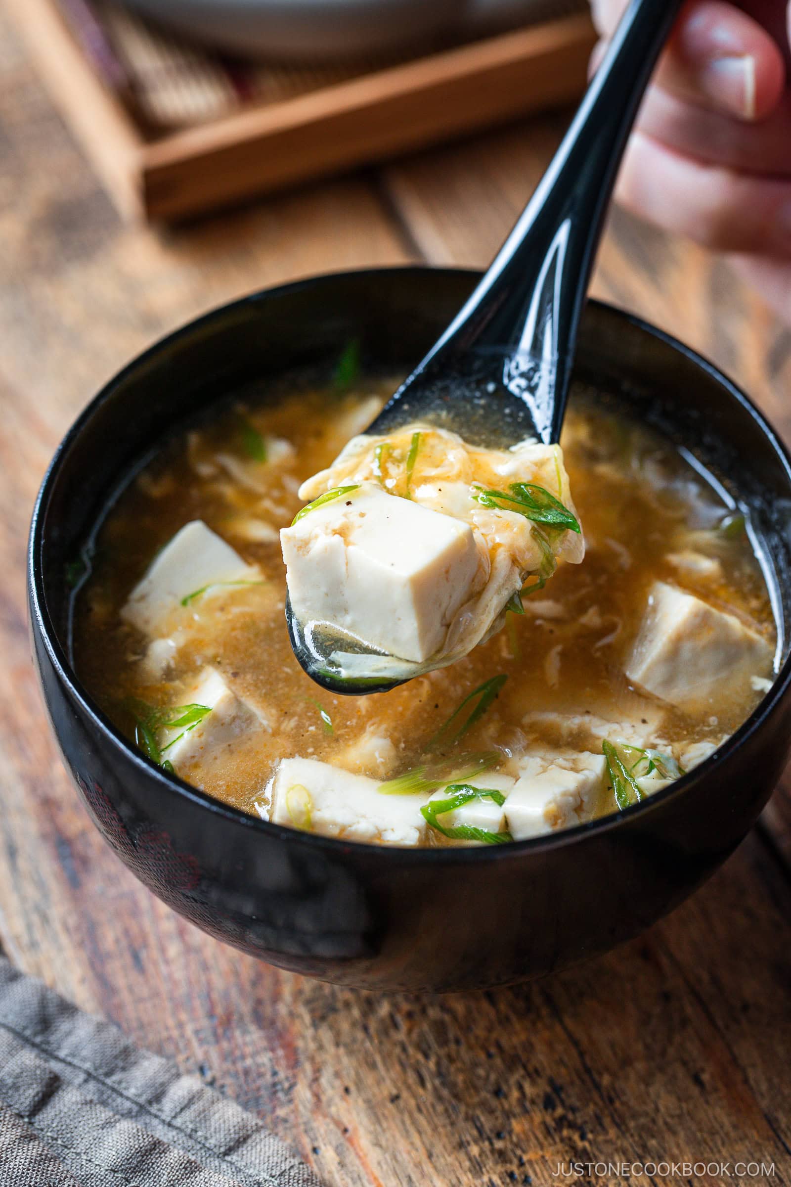 A black bowl of miso soup with tofu cubes and green onions, with a black spoon lifting tofu from the soup. The bowl is placed on a wooden surface.