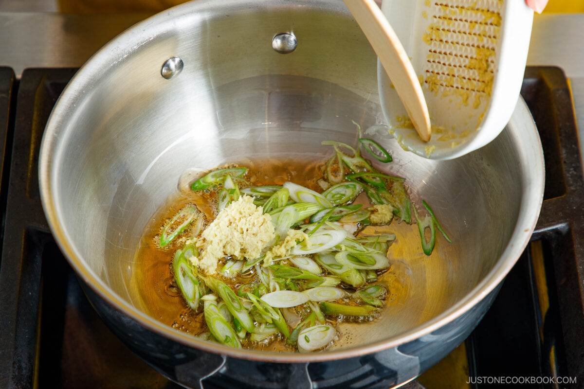 A hand adds grated ginger and sliced green onions to hot oil in a stainless steel pan on a stovetop. The mixture is being sautéed, and bubbles form around the ingredients.