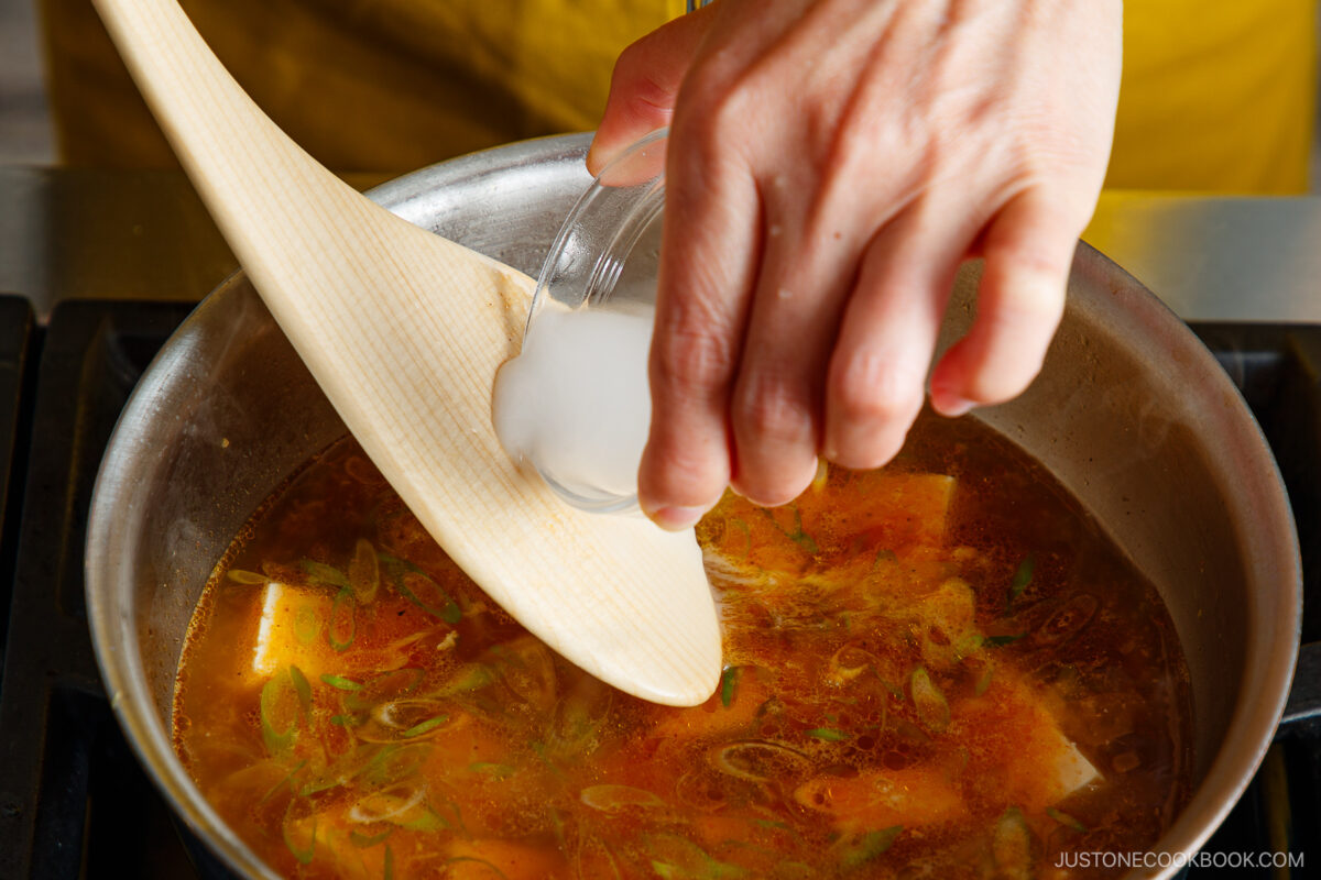 A hand pours a cornstarch slurry from a small glass bowl into a pot of simmering soup with tofu and green onions, using a wooden spatula to stir.