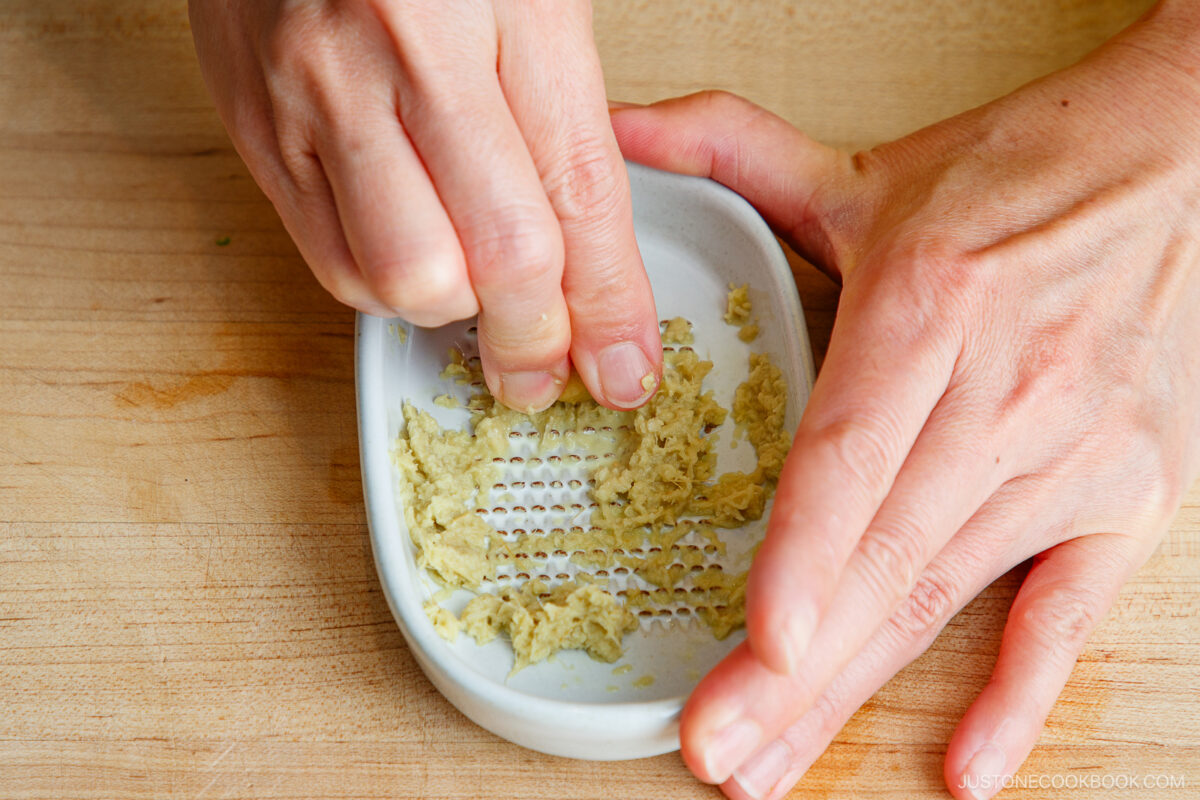 Two hands grating fresh ginger on a small white ceramic grater over a wooden surface. Grated ginger pieces are visible on the grater.