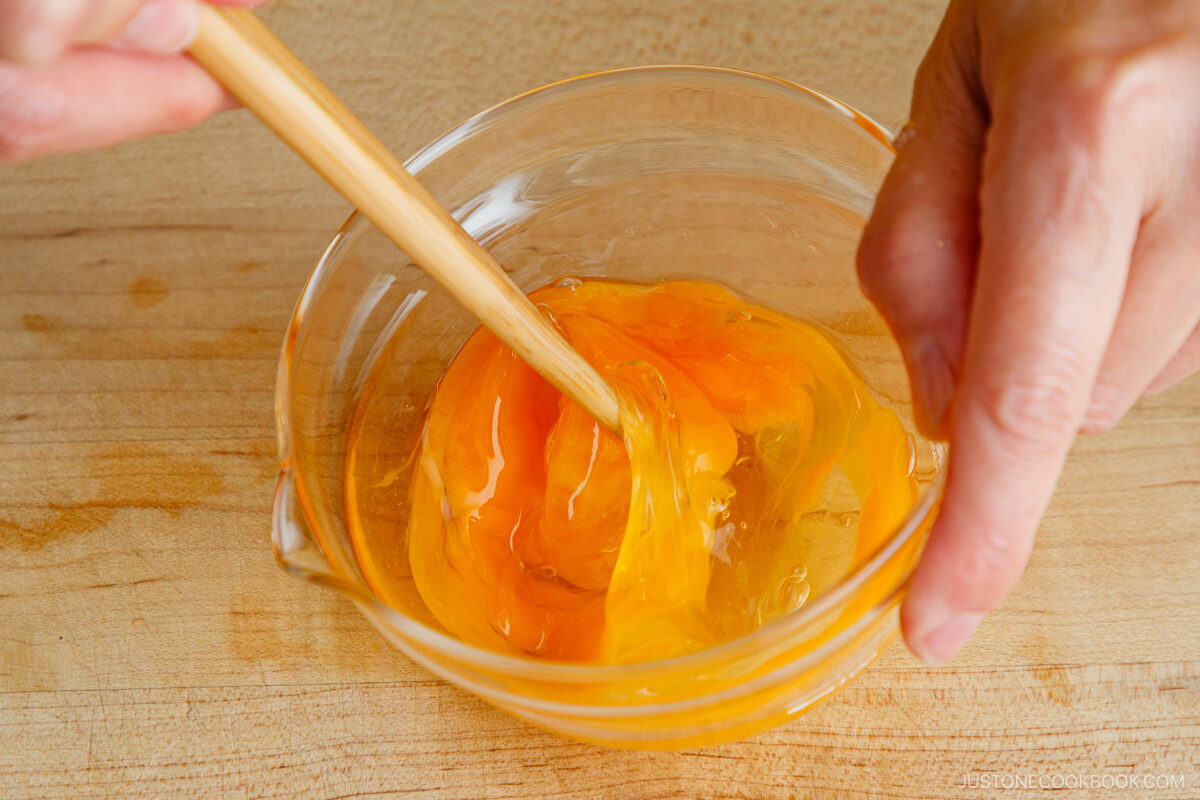 A close-up of hands using chopsticks to whisk raw eggs in a clear glass bowl on a wooden surface.
