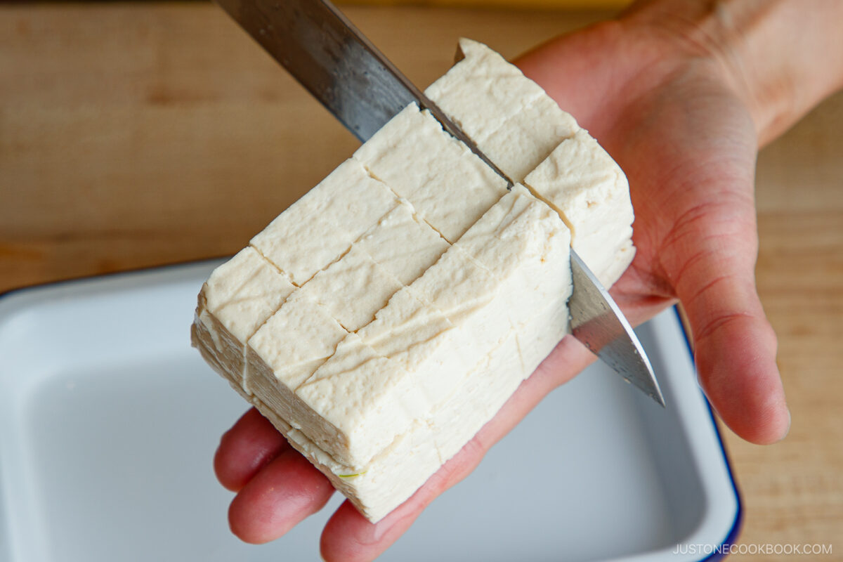 A hand holds a block of firm tofu while a knife cuts it into cubes over a white tray on a wooden surface.