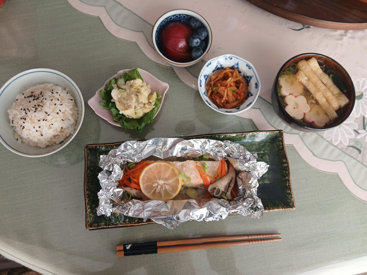 A Japanese meal with rice, miso soup, pickled vegetables, potato salad, fruit, and fish with lemon and vegetables in foil. Chopsticks are placed in front on a green tablecloth.
