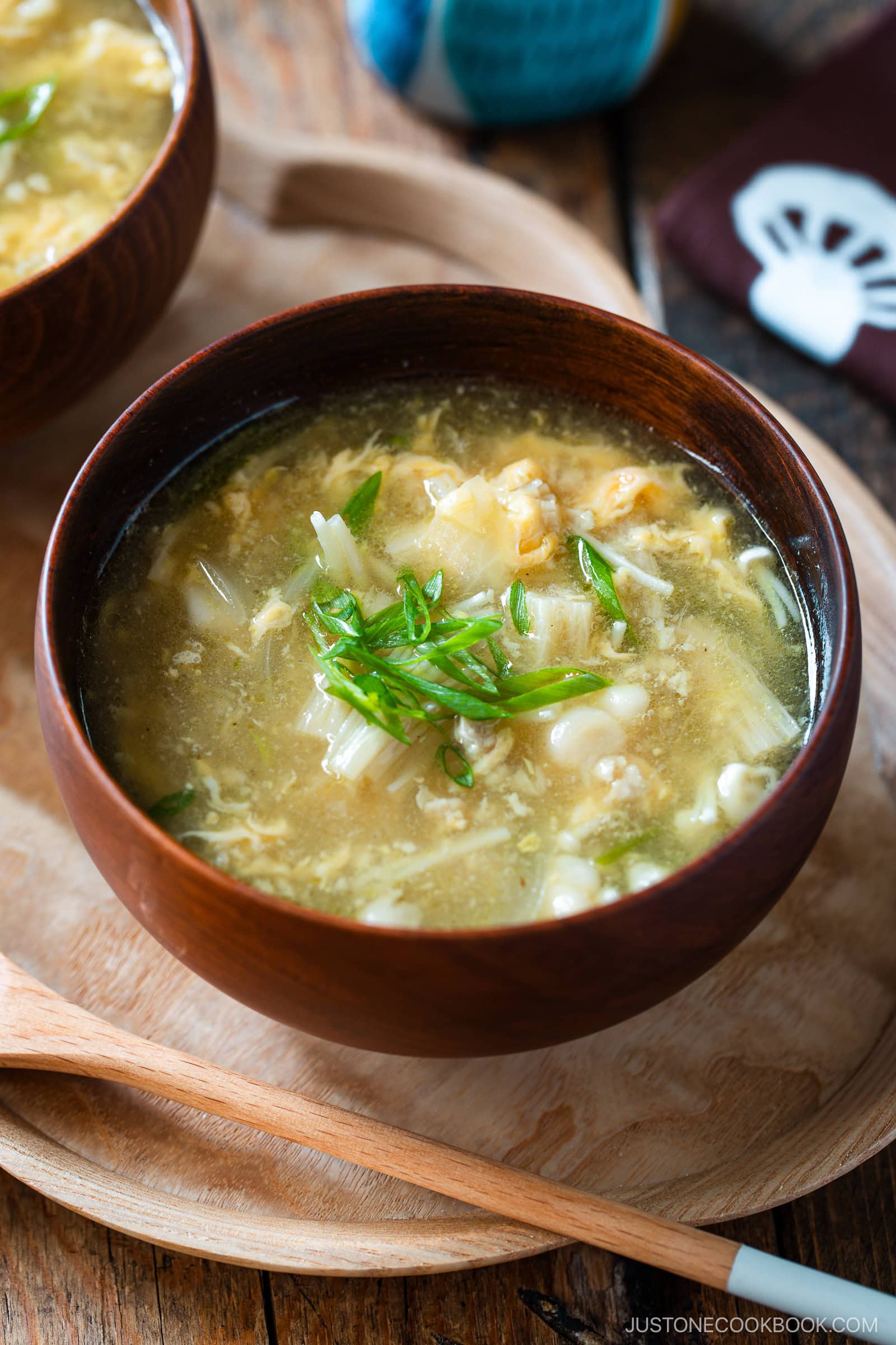 A wooden bowl filled with hot and sour soup, topped with chopped green onions, sits on a wooden tray with a wooden spoon beside it. Another bowl and a napkin are partially visible in the background.