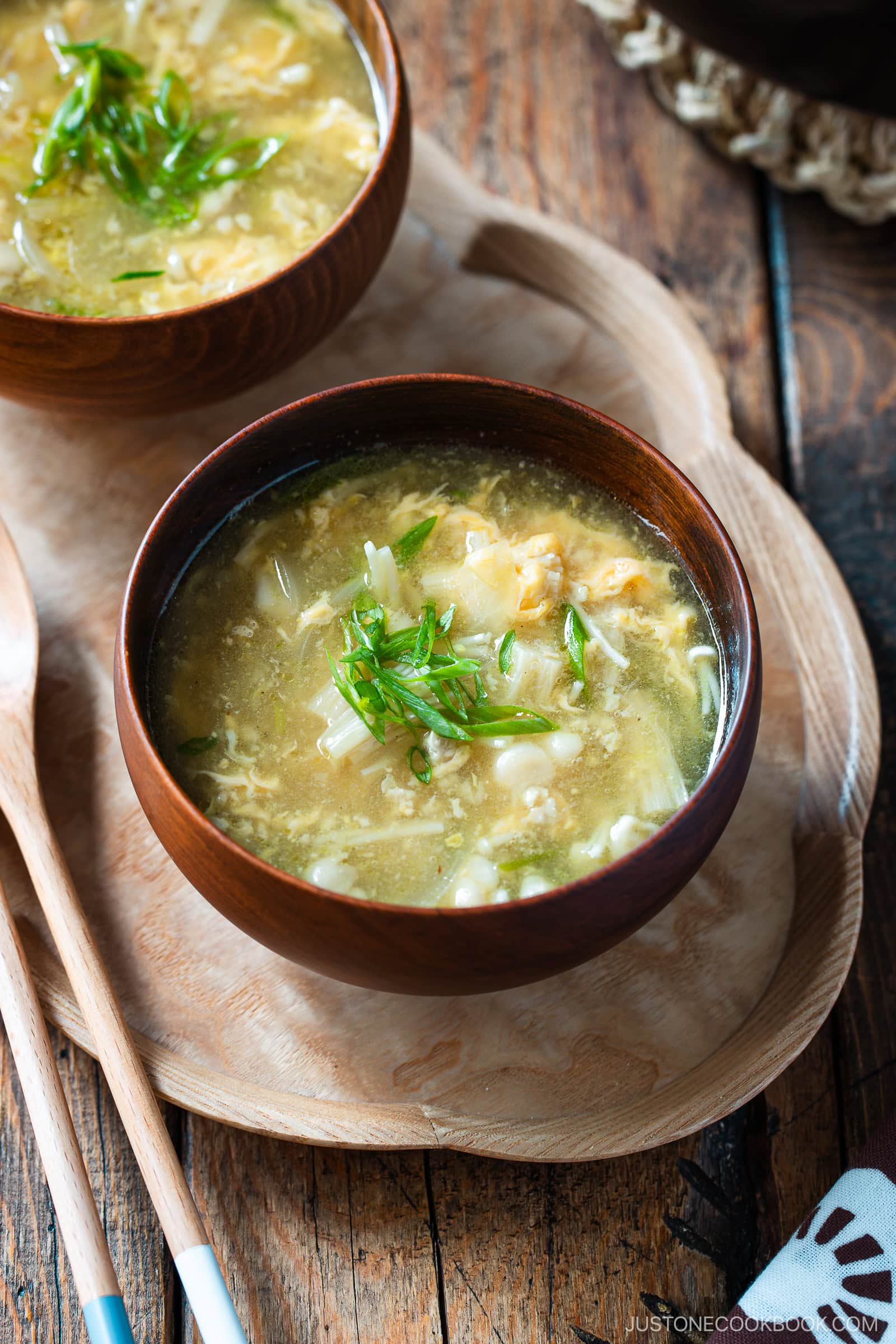 A wooden bowl of egg drop soup garnished with chopped green onions sits on a wooden tray, next to wooden spoons, with another bowl of soup partially visible in the background.