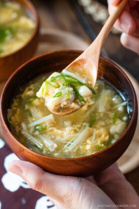 A hand holds a wooden bowl of clear soup filled with vegetables and tofu, while another hand lifts a spoonful of the soup, showing greens, tofu, and mushrooms.