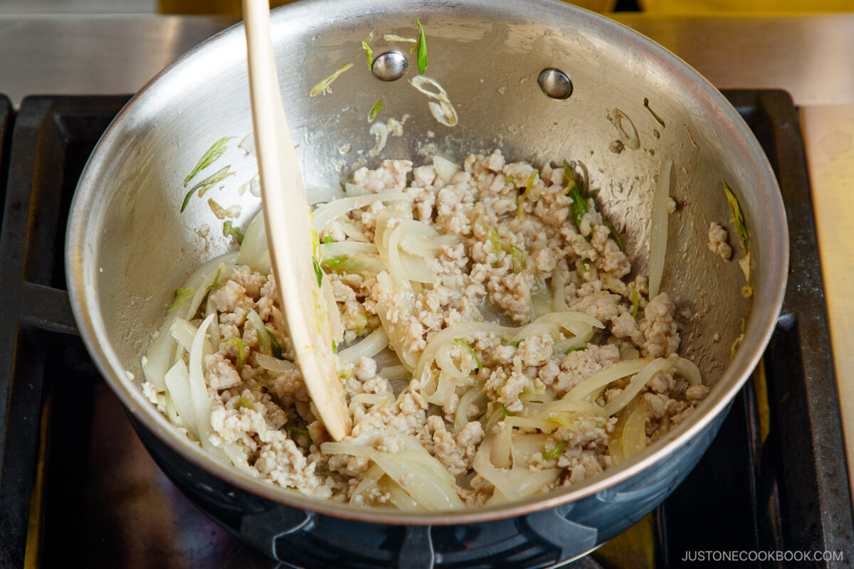 Ground meat, sliced onions, and green onions are being sautéed together in a stainless steel pan on a stovetop. A wooden spatula is stirring the mixture.