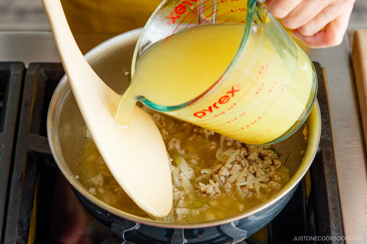 A hand pours yellow broth from a glass measuring cup into a pot of ground meat and onions on a stove, stirring with a wooden spoon.