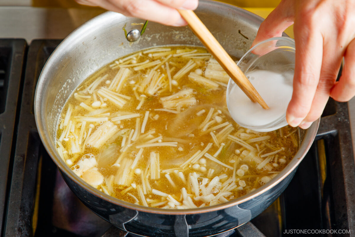 A hand pours a white liquid from a small bowl into a pot of soup with enoki mushrooms cooking on a stovetop. A wooden spoon is stirring the ingredients.