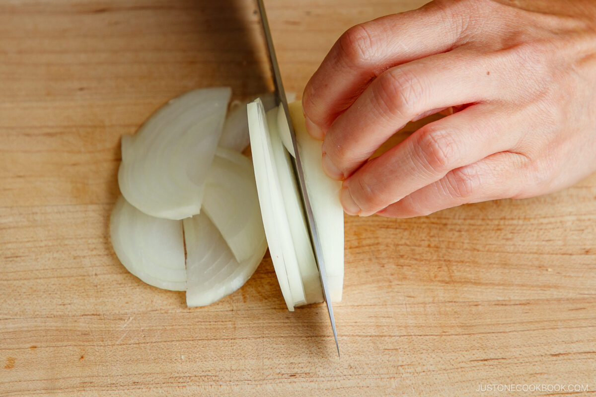 A hand slices a white onion on a wooden cutting board with a knife, creating thin, even rings of onion.