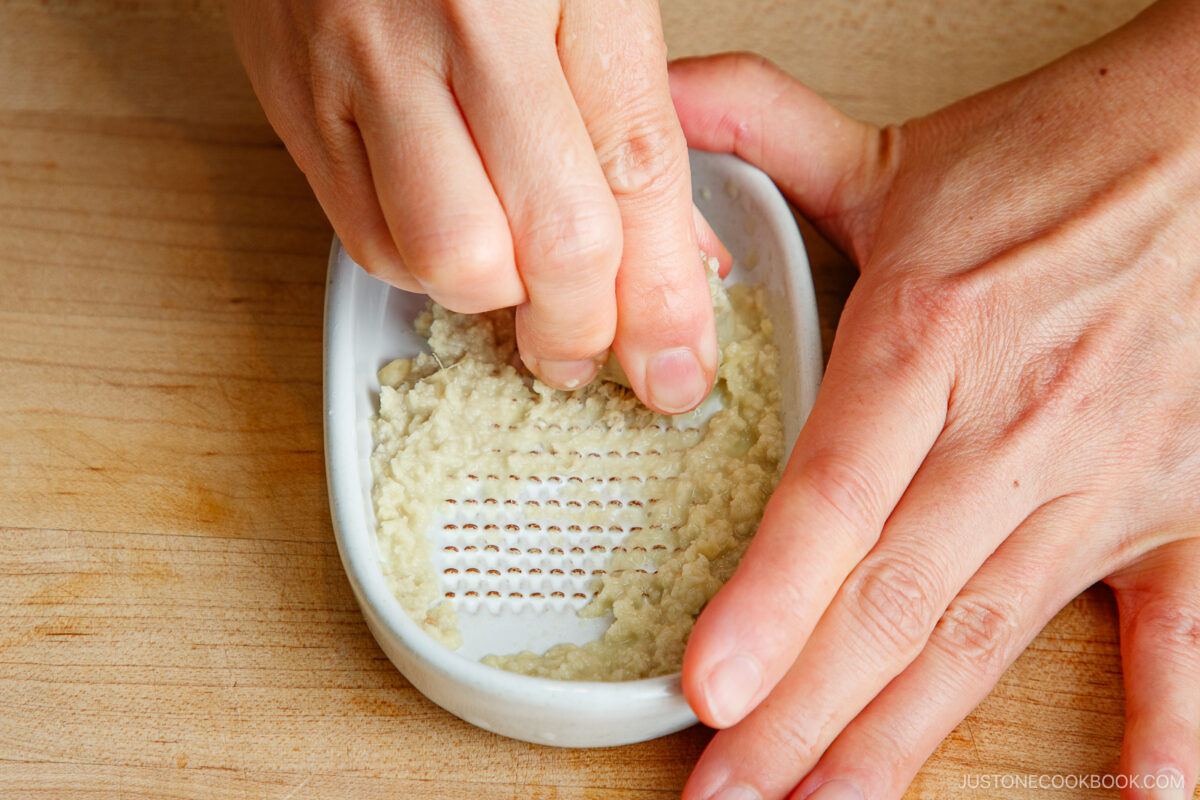 Close-up of hands grating ginger on a white ceramic grater, with grated ginger collecting in the dish, on a wooden surface.