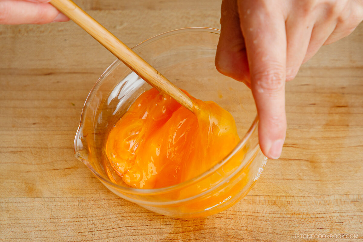 A close-up of hands using chopsticks to mix bright orange egg yolks in a glass measuring cup on a wooden surface.