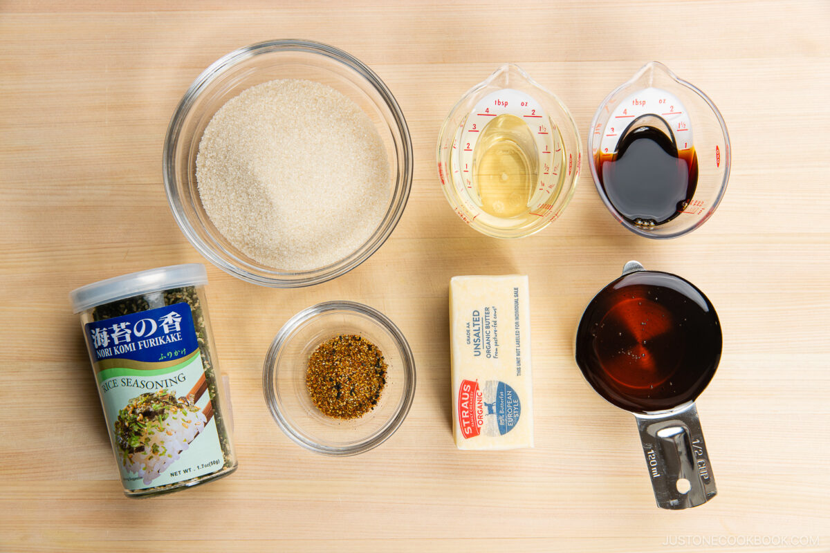 A top-down view of various ingredients in bowls and measuring cups on a wooden surface, including sugar, rice seasoning, spices, butter, vinegar, soy sauce, and a dark liquid.