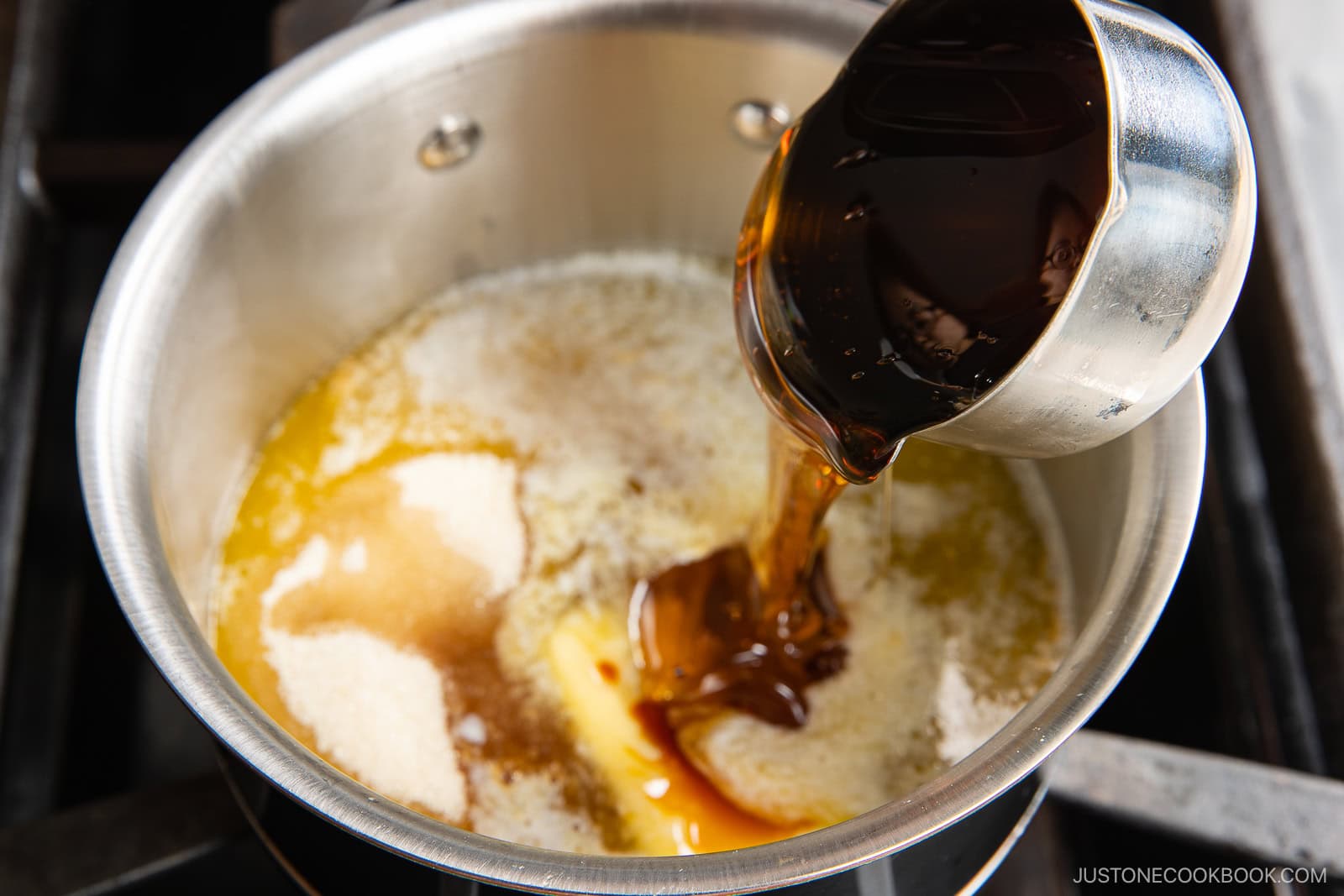 A metal measuring cup pours dark soy sauce into a saucepan containing melted butter and sugar, beginning the process of making a sauce on a stovetop.