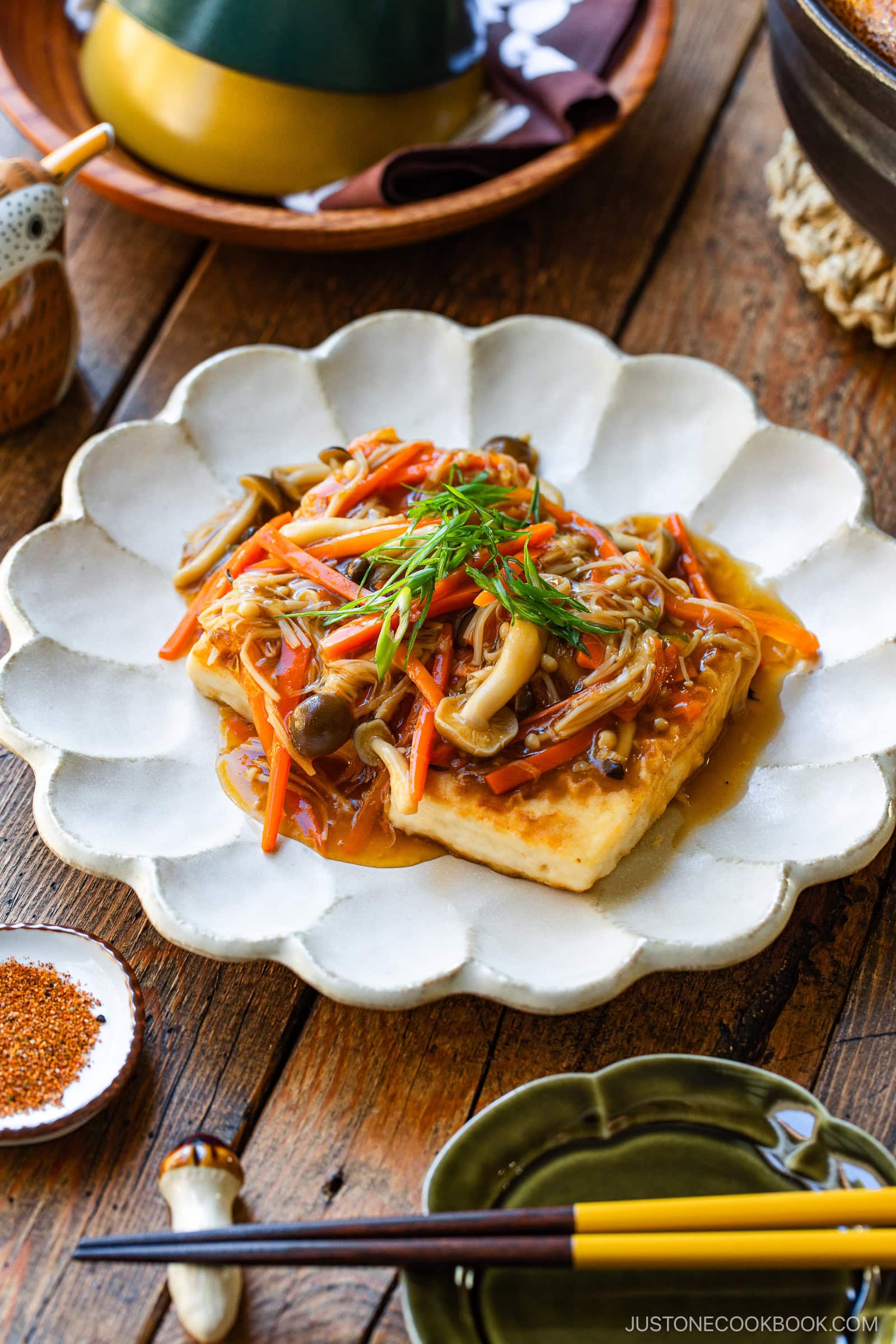 A plate of crispy tofu topped with mixed sautéed mushrooms, julienned carrots, and green onions in a savory sauce, served on a scalloped white dish on a rustic wooden table with chopsticks and condiments nearby.