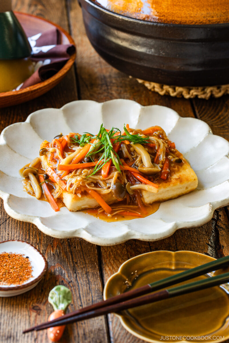 A plate of pan-fried tofu topped with a colorful vegetable stir-fry, including carrots and mushrooms, garnished with green onions. The dish is served on a rustic wooden table with chopsticks and condiments nearby.