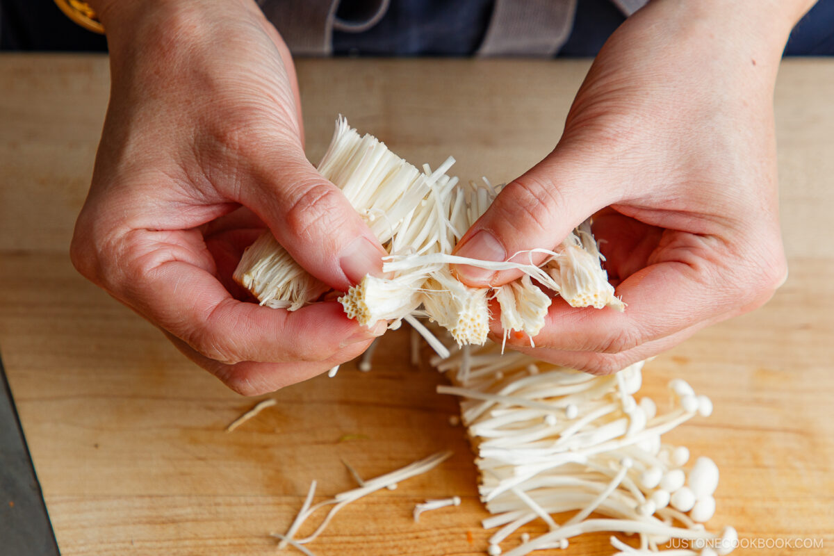 Close-up of hands separating a cluster of enoki mushrooms on a wooden cutting board.