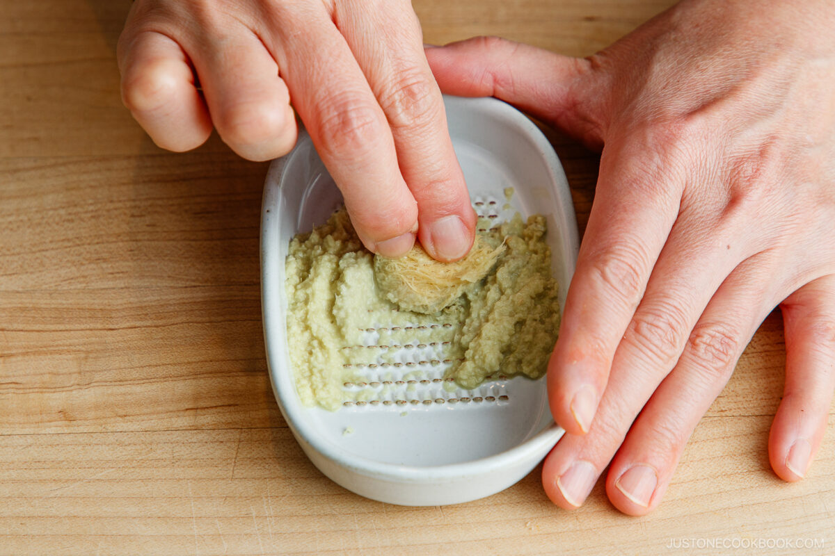 A person grating fresh wasabi root on a small white ceramic grater, with wasabi paste forming below, on a wooden surface.