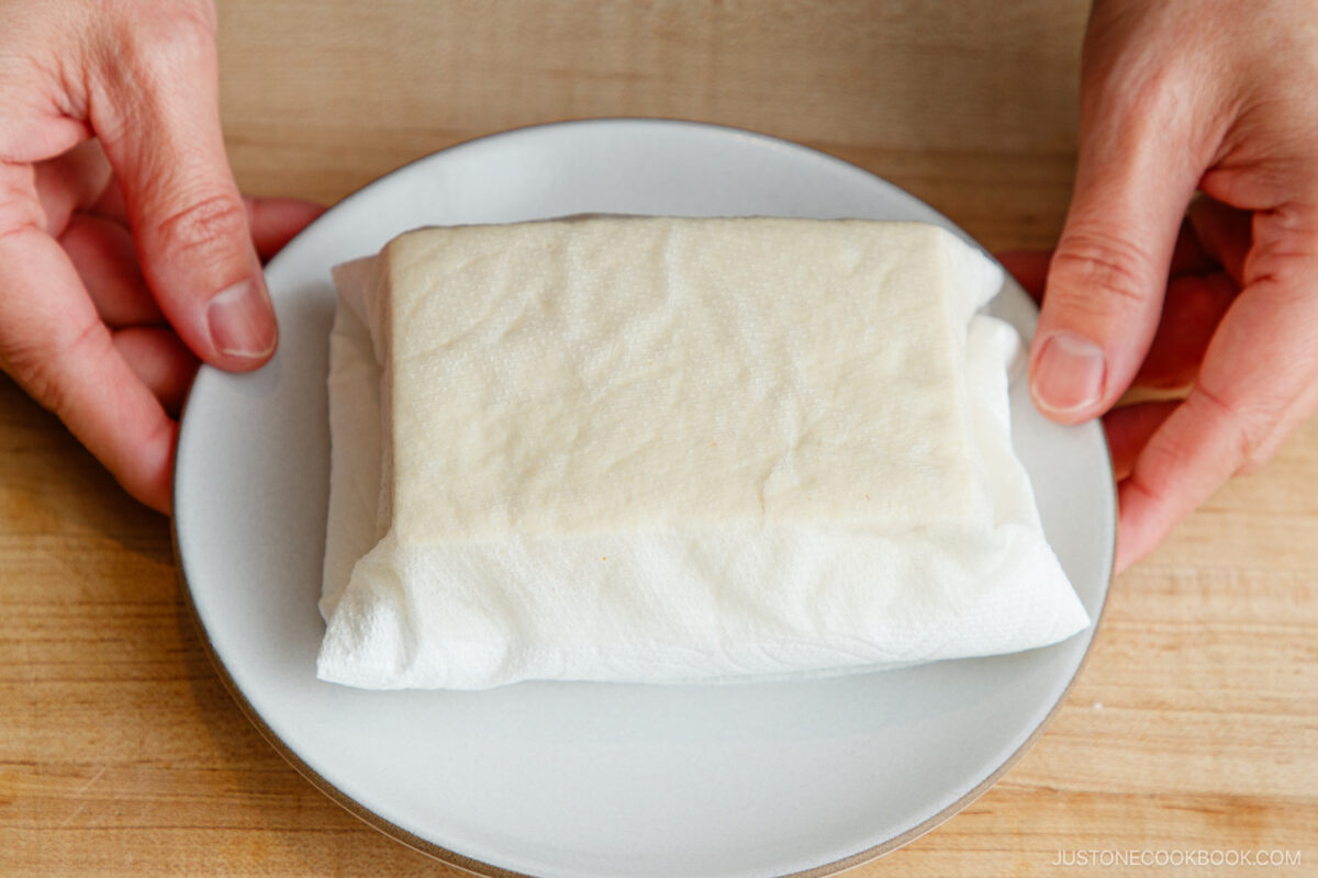 A block of tofu wrapped in a paper towel sits on a white plate, with two hands holding the plate on a wooden surface.